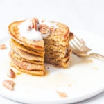 Stack of gluten-free blender pumpkin pancakes on a white plate with maple syrup and pecans.